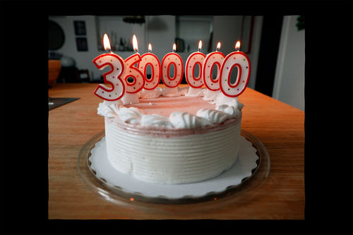 A white cake sits on a table with red candles that spell out 3600000 are lit on the top
