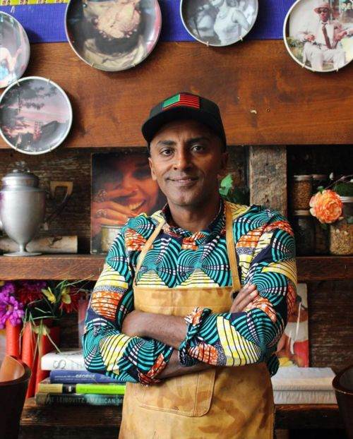 Smiling man in a colorful patterned shirt and tan apron stands arms crossed in a cozy shop, surrounded by dishes, produce, and rustic decor.