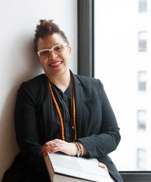 A woman with yellow framed glasses and curly picked up hair leaned againast a white wall, smiling and holding a large book