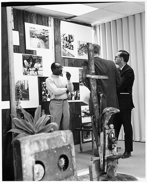 Noah Purifoy in front of a photographic installation of the Watts rebellion, by Harry Drinkwater, at the "66 Signs of Neon" exhibition at the University of Southern California, Los Angeles, c. 1966