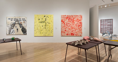 Foreground: tables from Mike Kelley, "Craft Morphology Flow Chart," 1991; on wall: three paintings by Sue Williams, from left to right: "The Art World Can Suck My Proverbial Dick," 1992, "Scooter," 1992, and "Record Profits," 2008; visible at right: works by Christopher Williams, Mary Kelly, and Tom Burr. Installation view at the Hammer Museum, Los Angeles
