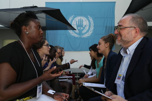 Two rows of adults sit facing one another, apparently in conversation. In the background on the wall hangs a light-blue flag with a logo on it that looks like an adaptation of the United Nations flag, with laurel leaves and an open hand, palm facing the viewer.