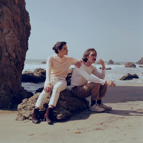 Pavo Pavo promotional image - woman and man sit on a rock on a rocky beach, with waves in the background.