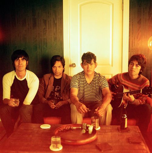 The Tracks band image - photograph of four figures sitting on a couch. The person on far right holds a guitar as if to play. A wood coffee table is in the foreground; in the background is a dark wall with a white door.
