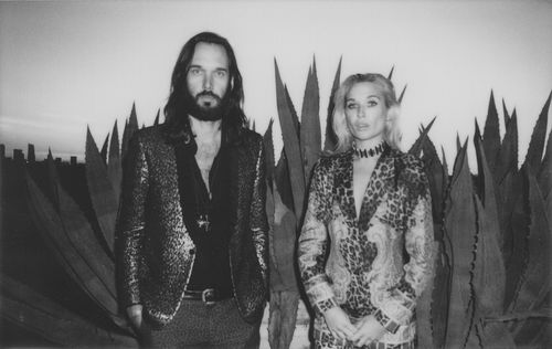 Wild Belle - black and white photo of a man and woman with tall cactus behind them and a city skyline at disk in the background. 