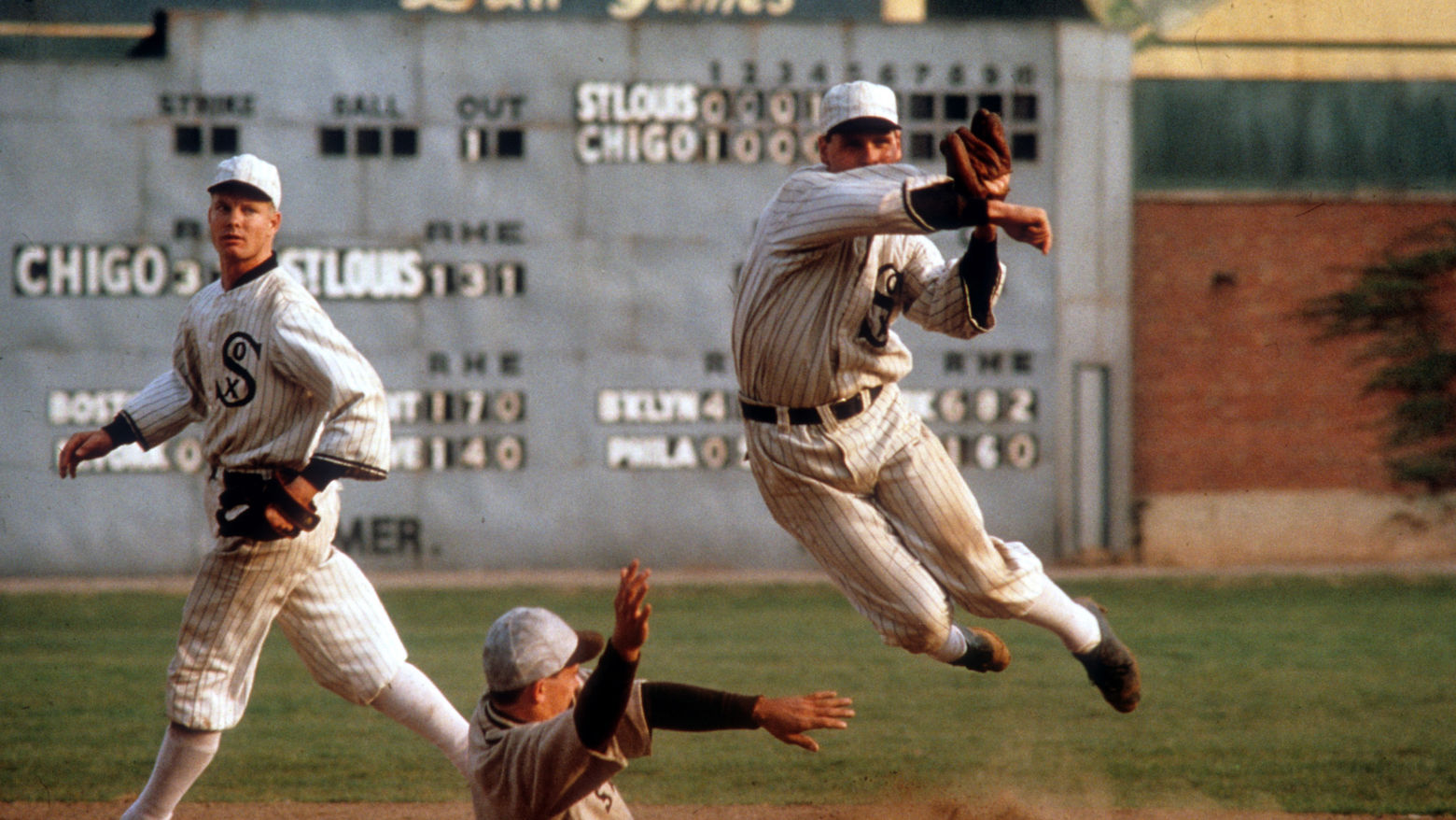 Still from the film Eight Men Out (1988) showing a baseball player slide into a base while another player, on the White Sox team, jumps and throws a ball to complete a double-play while a third player looks on