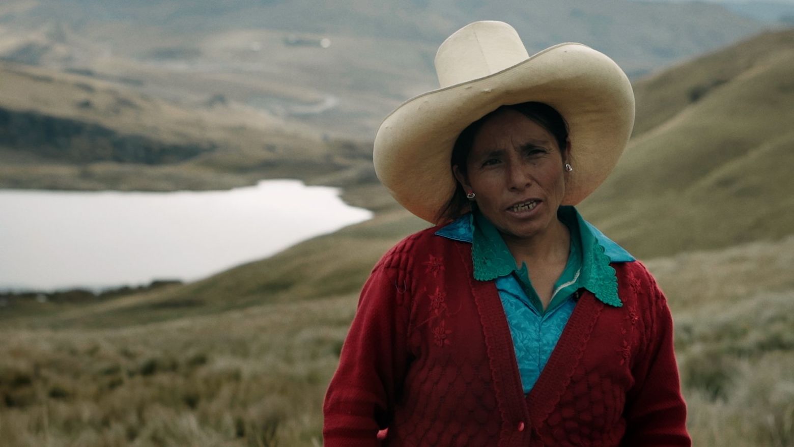 A woman stands outside against a hilly, green landscape with a lake or river in the background. The woman wears a large, white cowboy hat and wears a red cardigan over a blue shirt.