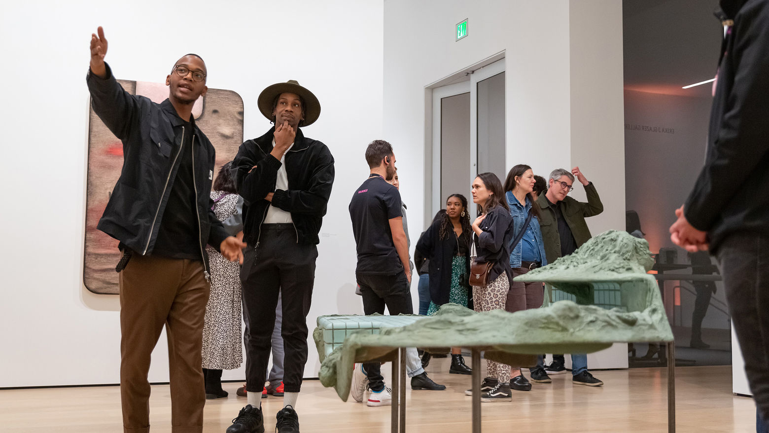 Two young man stand in a gallery next to a sculpture among a crowd