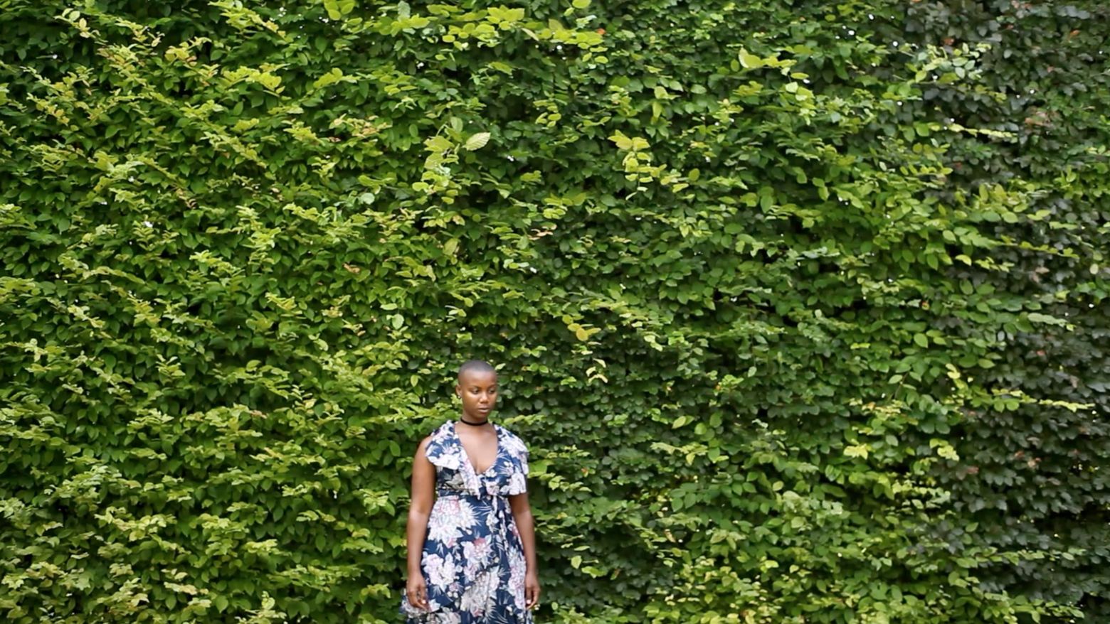 A woman in a dress stands facing the camera in front of a wall of plants