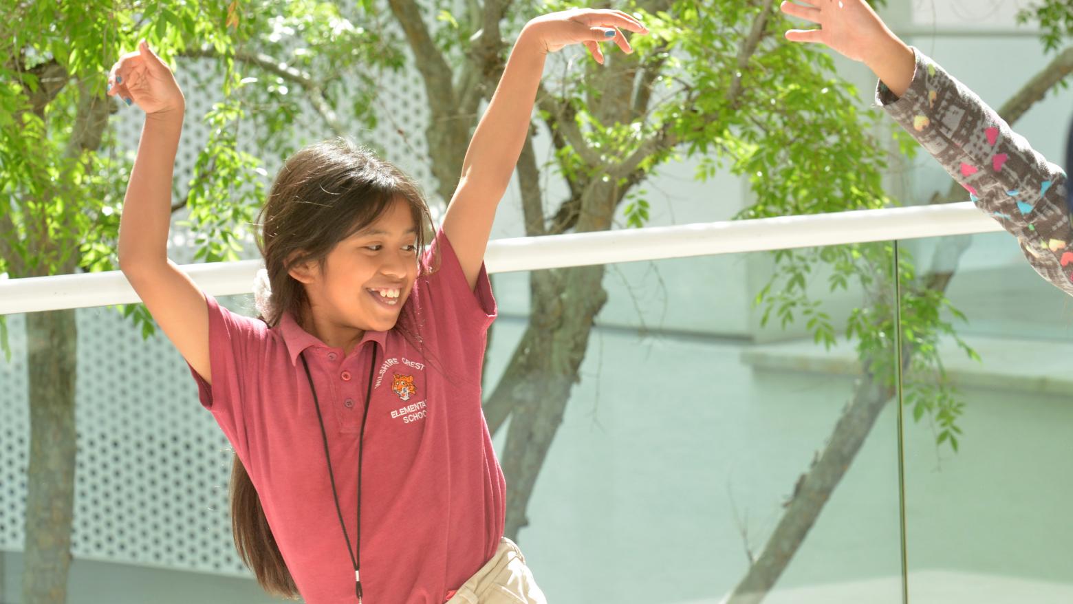 A young girl smiles while standing and raising her arms, and looking to her left at another person whose arm is visible in the photo.