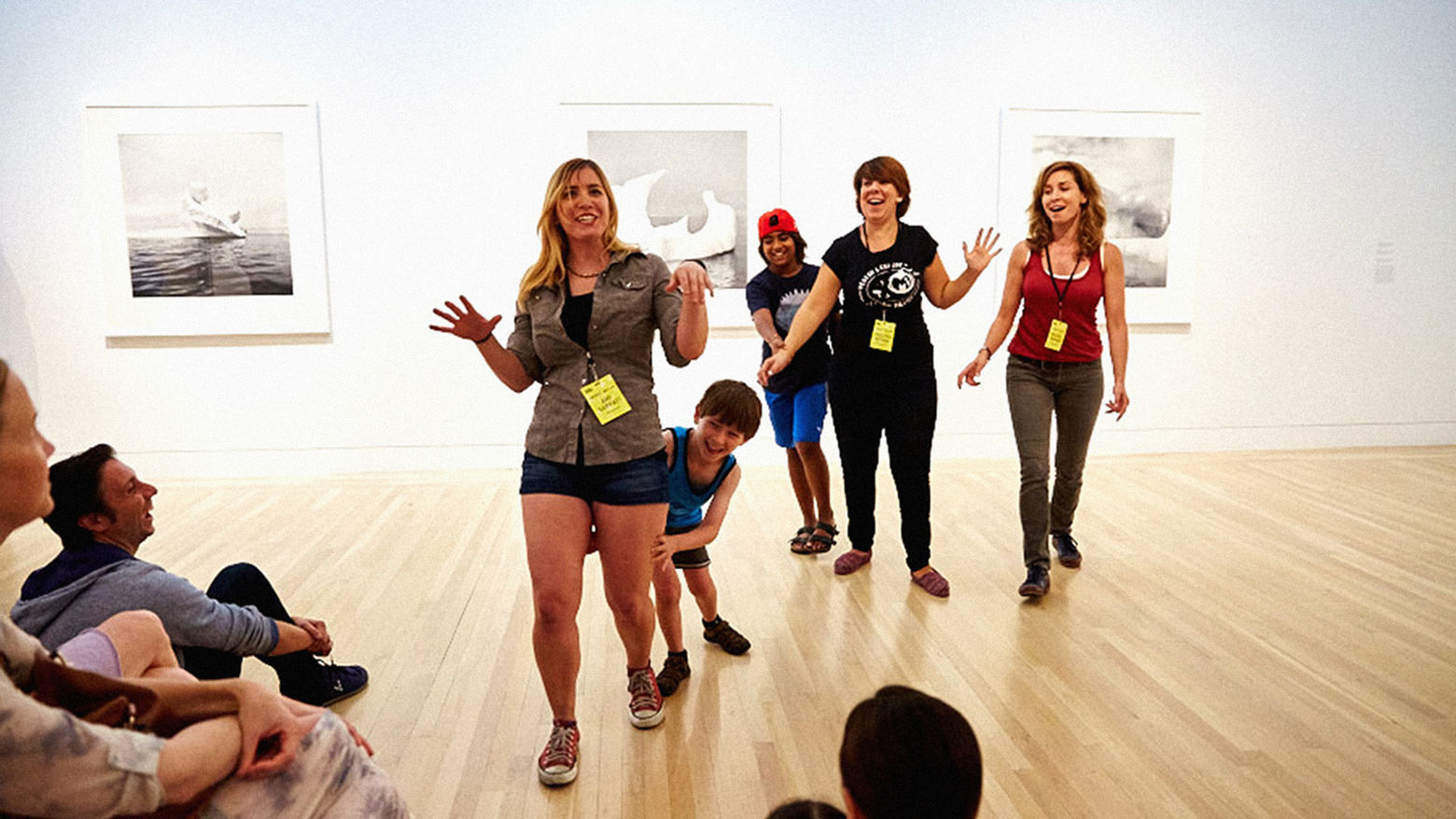 Children and adults in an art gallery, smiling and interacting. What appear to be black and white photographs are hanging on the wall.