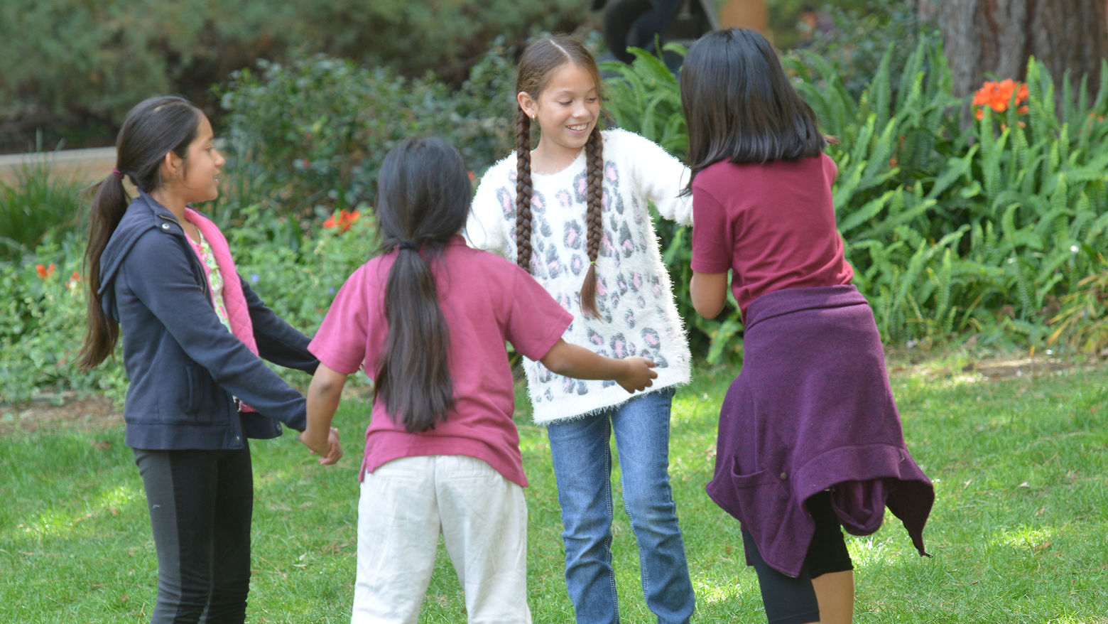 Children stand on a grassy area in a circle, holding hands and smiling, with plants in the background.