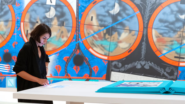 Visitor in a gallery stands, looking down at a device embedded in a table. A very large teal book is also on the table. In the background is a wall-sized painting with geometric designs, red and blue background with black lines.circles,