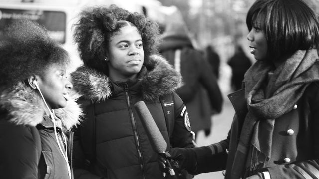 Two young women in coats face a woman holding a microphone