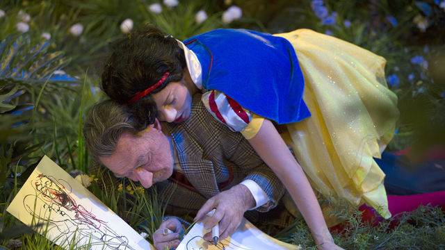 A figure dressed as Snow White lies on top of a man drawing in the grass