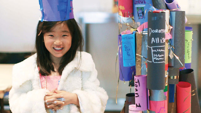 A young girl stands, smiling, wearing a hat made of blue construction paper, a white coat, and silver glitter on her fingers. To the right of her is a structure made of rolls of colored construction paper with words and tinsel.