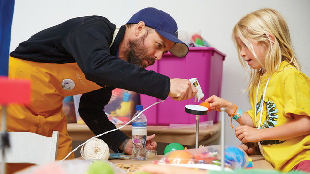 A girl in a yellow shirt is holding up an art project and is assisted by a man in a baseball cap who leans over with a glue gun pointed at the girl's project.
