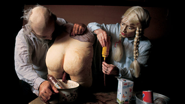 Two people wearing doll-like masks lean over a table where a nude bottom that appears made from plaster or papier-mache is sitting, Various bowls and pots sit on the table as well.