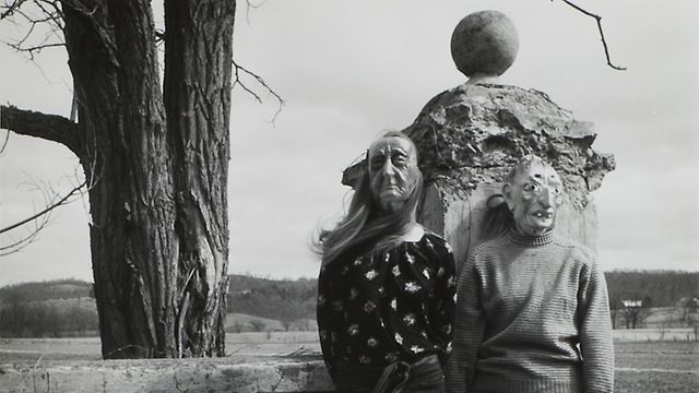 Two figures in masks stand before a stone wall