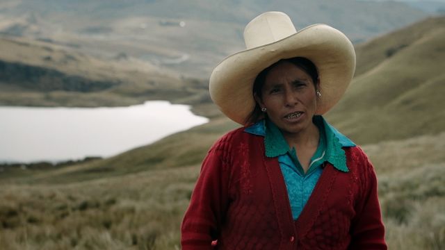 A woman stands outside against a hilly, green landscape with a lake or river in the background. The woman wears a large, white cowboy hat and wears a red cardigan over a blue shirt.