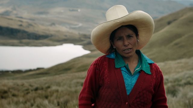 A woman stands outside against a hilly, green landscape with a lake  or river in the background. The woman wears a large, white cowboy hat and wears a red cardigan over a blue shirt.