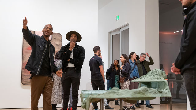 Two young man stand in a gallery next to a sculpture among a crowd