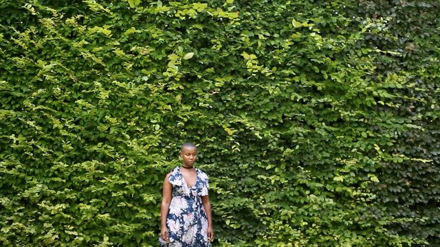 A woman in a dress stands facing the camera in front of a wall of plants