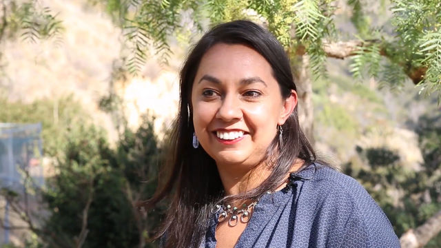 Photo of artist Mercedes Dorame, a woman with long brown hair smiling at someone standing to left of cameral. She is outside, with a hillside of foliage behind her. She wears a blue shirt and a silver necklace.
