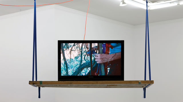 An open laptop computer sits on a wooden shelf that hangs from the ceiling. The walls in the background are white, and the edge of an open skylight are visible at the top right of the image.  