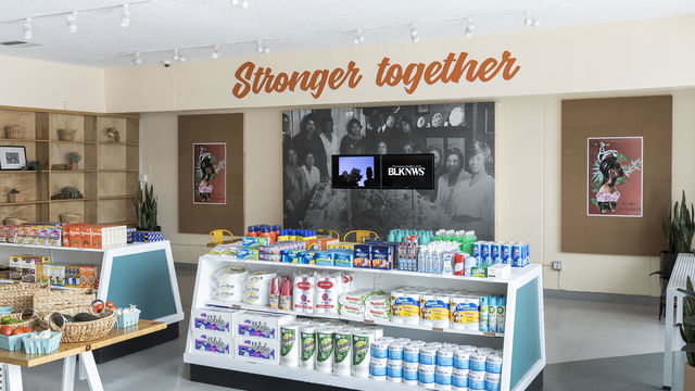 Photograph showing inside of a grocery store, with low shelving. On the back wall the words "Stronger Together" are written in orange cursive. Below this a black and white photographic mural, with two video monitors mounted over top.