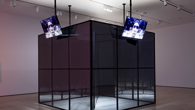 A large, mirror-like cube in the middle of a gallery, with television screens along each side