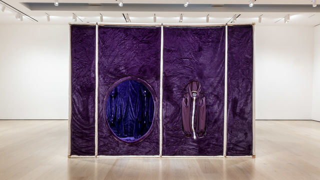 A large gallery with white walls. In the center of the room a large square structure of white poles and purple fabric covering the surface. On the  side of the structure there is a large hole, which provides a view of the inside.