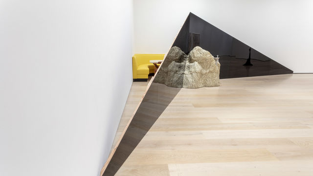 A view of the corner of a gallery. Two dark triangular structures extend across the corner, meeting in the middle at the tall ends. A large rock sits on the floor in front of the triangles, where they meet. Behind the triangles, between them and the wall, a yellow corner booth and a table are visible.