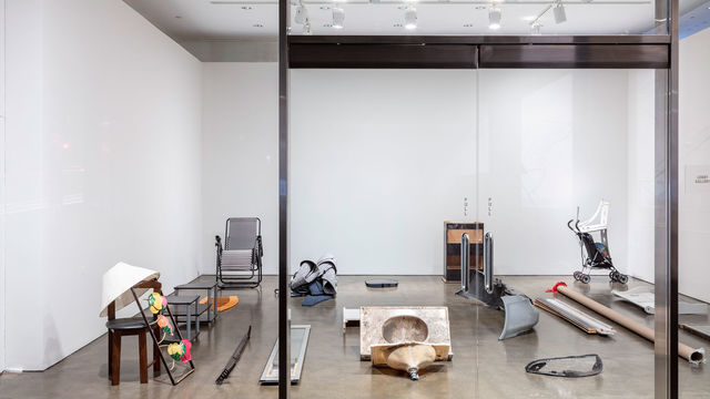 View looking into a room through a glass wall and glass doors. The room has various objects sitting on the floor, such as a lamp, a sink, a chair, blankets.