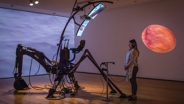 A person stands in a dimly lit gallery with abstract video projections on the walls, wearing headphones connected to a mechanical sculptural installation by the artist Sondra Perry