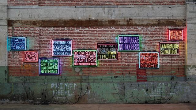 Photo of neon light artworks installed on a brick wall. The neon artworks are signs with phrases in Spanish and English related to immigration.