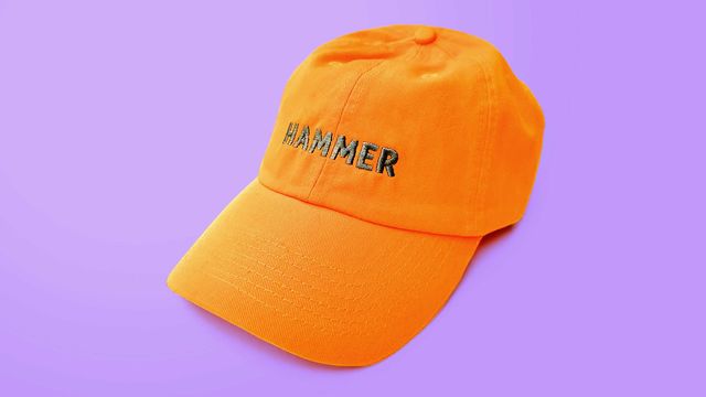 A bright orange baseball hat sits on a uniform lilac background. The had is embroidered in the forehead with the word HAMMER in dark grey .