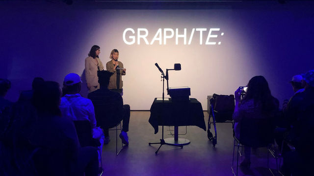 Photo of a dark room with the farr wall spotlit and the word "GRAPHITE:" projected on the wal in white letters. On the left and right we see the backs of people sitting in chairs. Two women stand against the far wall and speak into a microphone. 