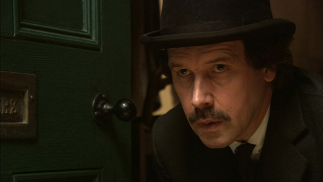 Still from the film "Bloom" (2003) showing actor Stephen Rea portraying the character Bloom, wearing a black bowler hat and peering through a doorway.