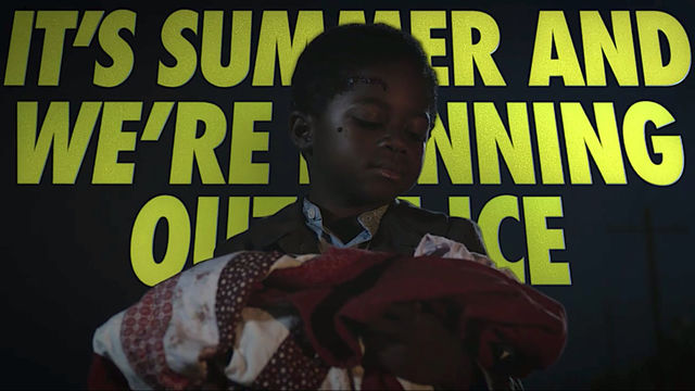 Still from the HBO television series "Watchmen," showing a young black boy holding a baby wrapped in a blanket, superimposed over large yellow type that reads "IT'S SUMMER AND WE ARE RUNNING OUT OF ICE"