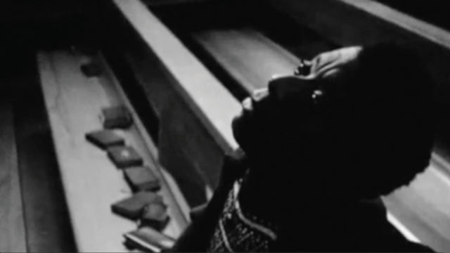 Black and white photograph, with a person in the foreground, visible from the chest up, sitting in a church pew and looking upwards. The person has dark skin and short dark hair. The pew extends diagonally into background at the upper left corner. Books are strewn on the pew next to the person. 