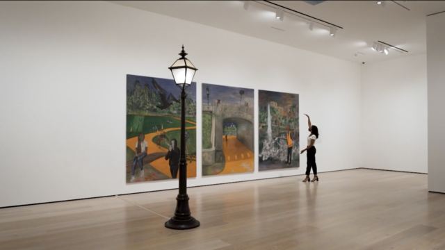 Lightpole in front of a triptych. One woman stands to the right of the paintings with one arm raised