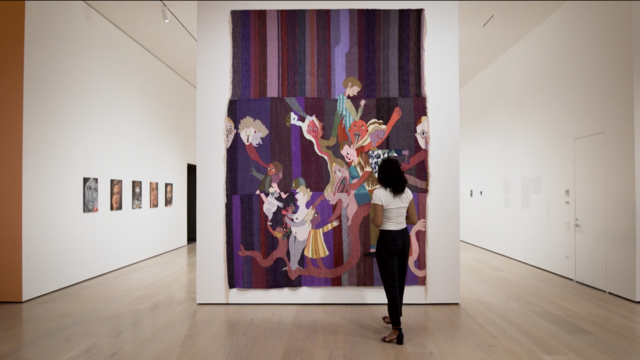 A woman stands center right in front of a multi-colored tapestry with a purple background and many human-like figures in the foreground