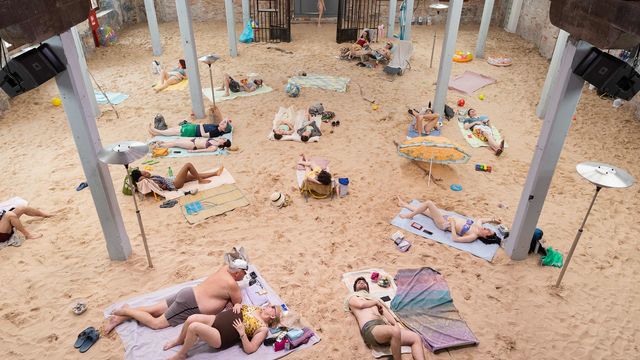 Photograph of people in bathing suits laying on a sandy ground inside of a building with columns.