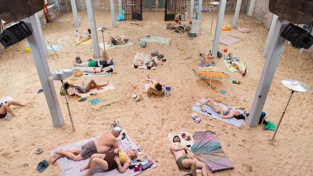 Photograph of people in bathing suits laying on a sandy ground inside of a building with columns. 