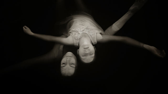 Black and white image with a black background. Two figures emerge from the dark, as if lying backwards, with heads hanging upside down. The figures have light skin and dark hair and have their eyes closed and arms outstretched.