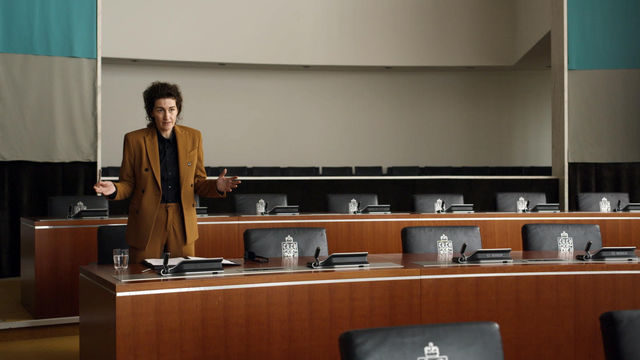 IMage of a woman standing in a meeting room with rows of wooden rows of large chairs.  