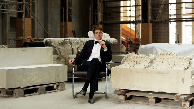 A light-skinned person with short dark hair, sits on a modern chair, wearing a tuxedo  inside a wearhouse with large stone scultuptural objects on pallettes.