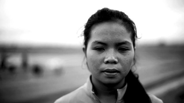 Black and white image of a person seen from the shoulders up and facing the camera, but eyes turned down. The background is a blurred image of a landscape with diagobal lines from left to right.  