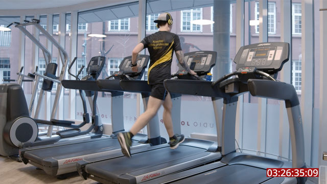 IMage of a person wearing black shorts and sturt and a baseball cap, running on a treadmill in a room with other treadmills and  large windows in the dar wall. 